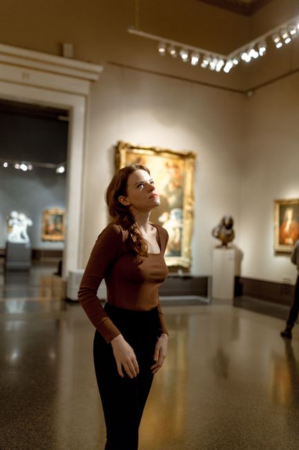 Caucasian woman in brown sweater gazes at artwork in an art museum gallery.