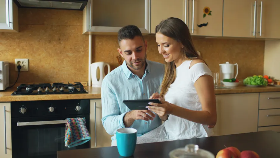 Happy couple using a tablet in a cozy kitchen setting, discussing a recipe.
