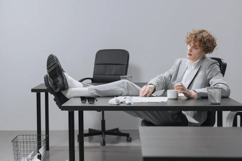 A curly-haired man in a suit relaxes with feet on desk, looking serious in a modern office setting.