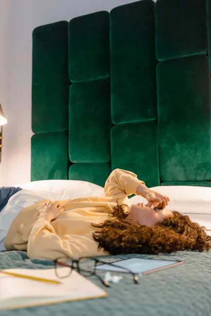 A woman lies down in a bedroom, looking tired with a headache, surrounded by work items.