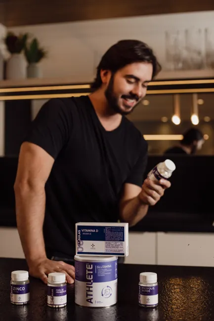 Adult man standing in a kitchen examining a vitamin bottle with more supplements on the countertop.