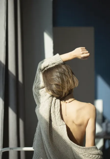 Back view of a woman in a knitted sweater enjoying sunlight indoors.