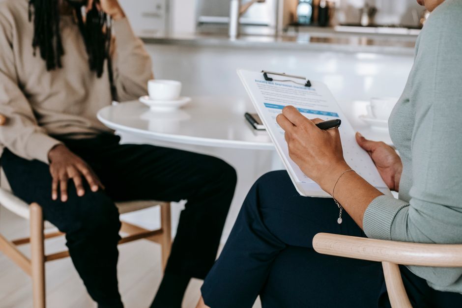 Crop unrecognizable female psychotherapist or interviewer asking questions and taking notes in clipboard during interview with black male
