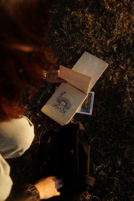 A person observes a detailed sketch in a notepad on grass under warm, natural lighting.