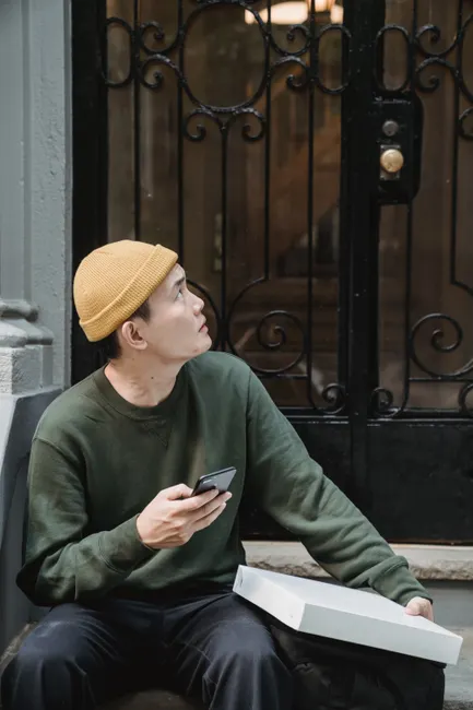 A puzzled delivery man sits outdoors, holding a phone and box, looking up at a door.