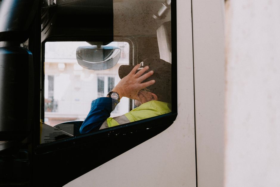 A truck driver in neon jacket rests with hand on head inside vehicle cabin, view from the side window.