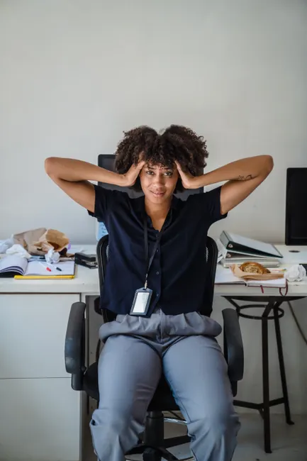 A worried office worker sits at a cluttered desk, overwhelmed by paperwork.