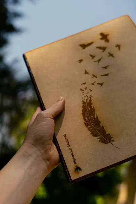 Close-up of a hand holding a notebook featuring an elegant feather and bird design in natural outdoor setting.