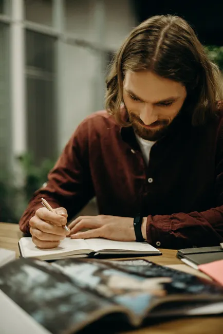 Man writing in a notebook, focused on his work, surrounded by books and papers.