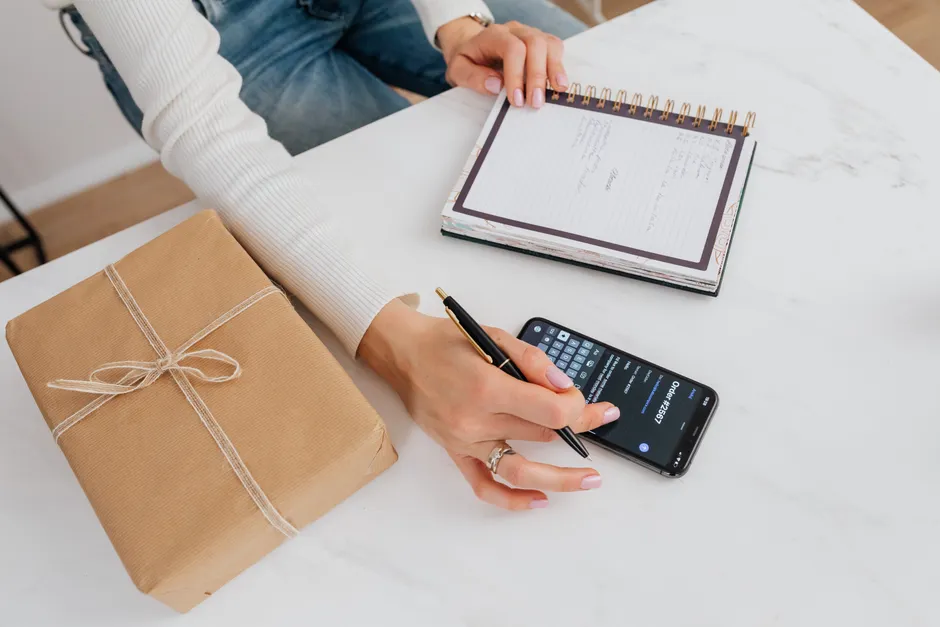 Person writing notes in a notebook beside a smartphone on a desk with a wrapped gift.