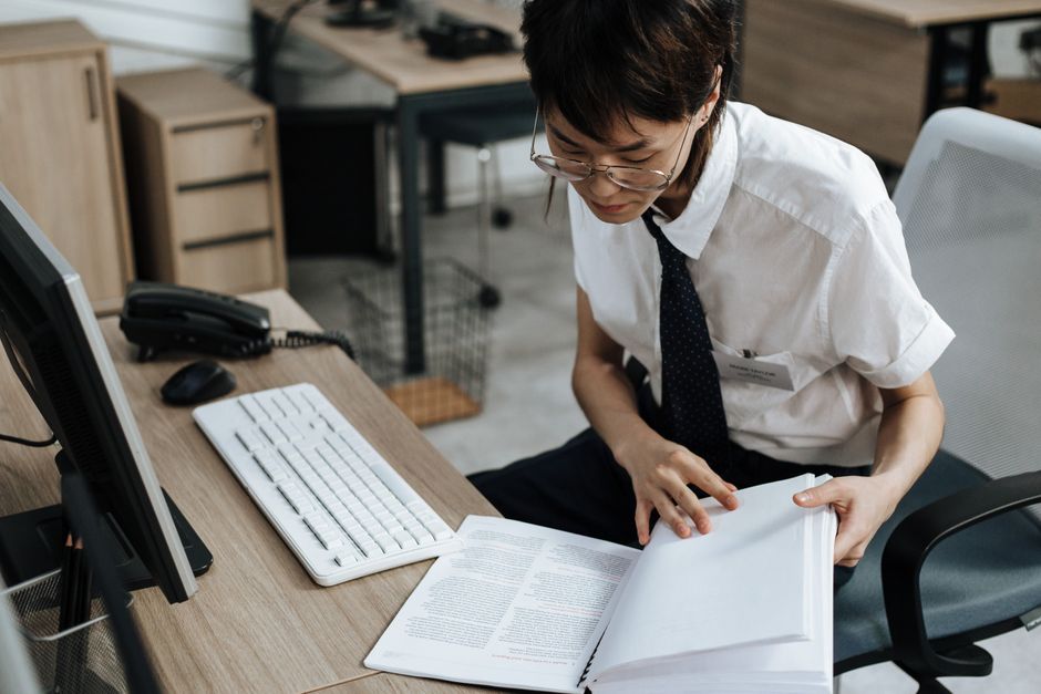 Professional in office attire reviewing documents at a desk with computer and book.