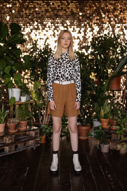 Young woman wearing leopard print standing among potted plants indoors against a sparkling backdrop.